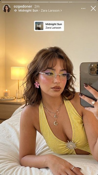 A woman with rhinestone glasses takes a golden selfie on white bedding