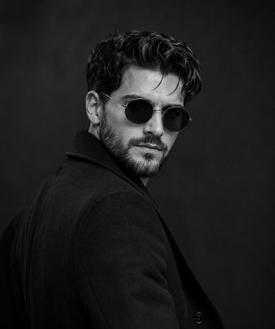 A confident man in black overcoat turns with intense gaze