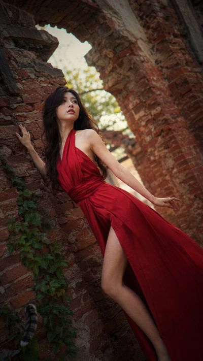 East Asian woman in crimson silk gown poses beneath crumbling brick arch