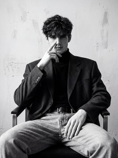 Curly-haired man in plaid blazer sits in dramatic monochrome portrait