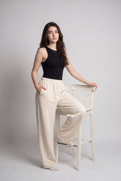 Model in black tank and cream pants poses with one leg on chair