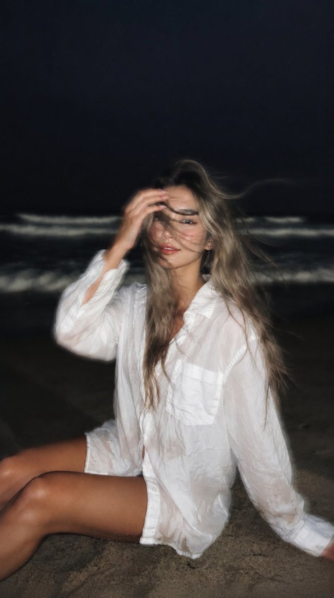 Girl sits on dark sand as wind blows her wet hair across her face