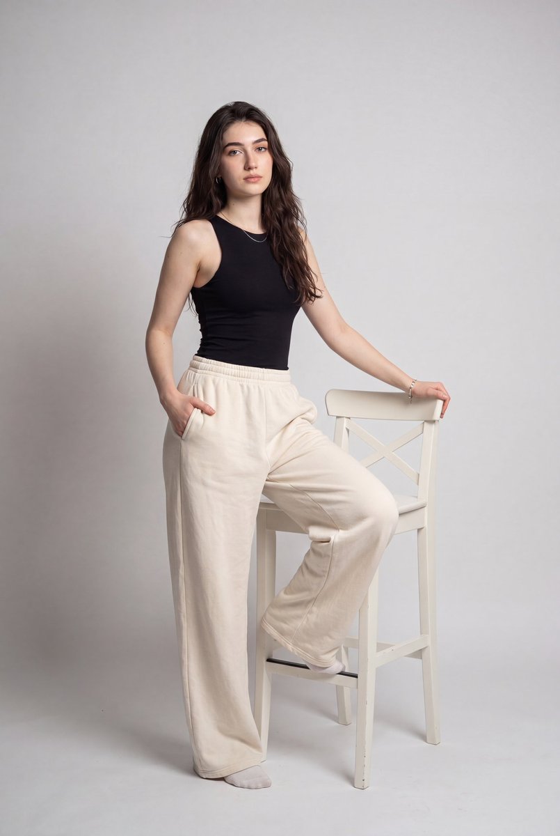Model in black tank and cream pants poses with one leg on chair