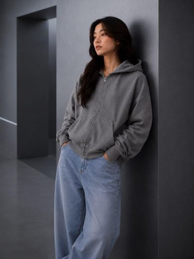 Young woman in oversized hoodie leans against gray wall in soft light