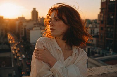A melancholic woman with short red hair stands on a rooftop at golden hour
