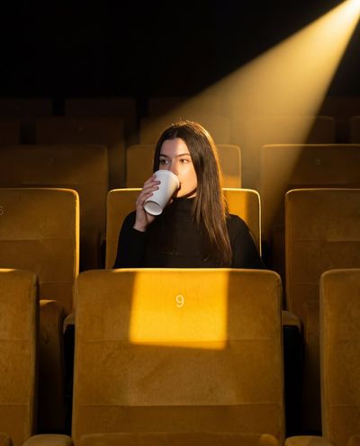 A solitary woman in a dark theater sips from a glowing cup