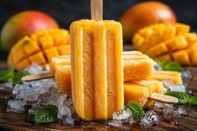 A vertical mango popsicle sparkles with frost on a dark wooden board