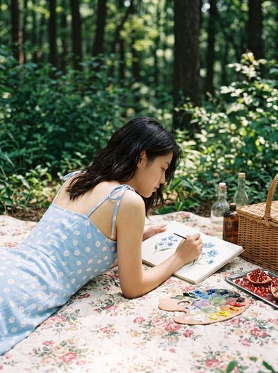 Woman painting on picnic quilt in sunlit forest clearing