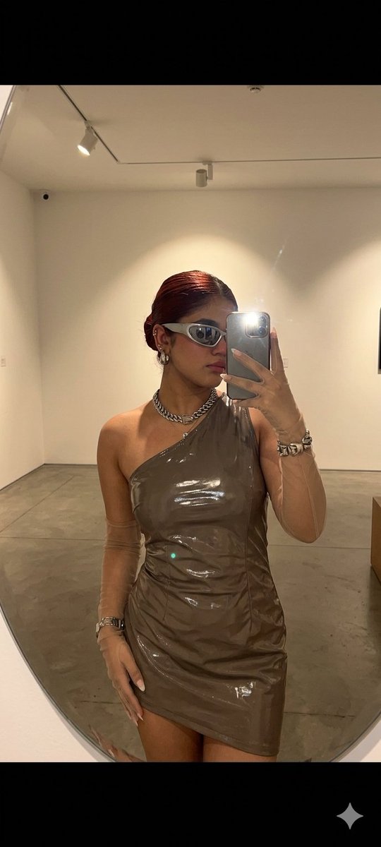 A woman in a futuristic dress takes a mirror selfie in a gallery.