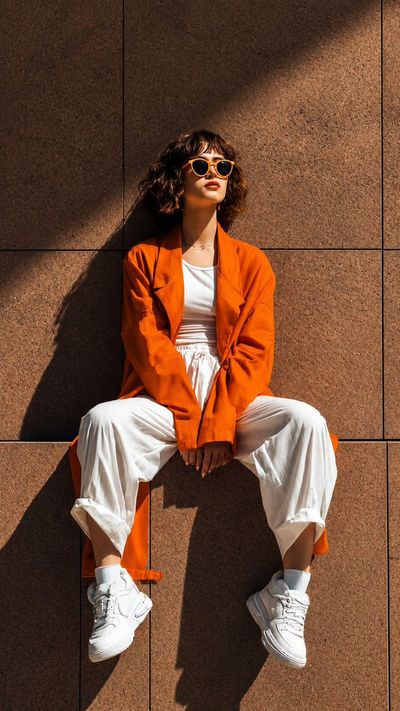 A confident model sits against a stone wall in orange coat.