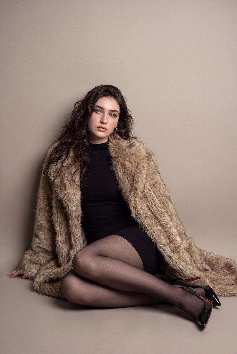A model sits on the floor in a warm studio wearing a fur coat.