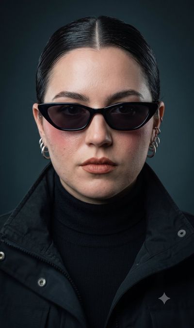 A woman in black sunglasses stares intensely under studio lights.