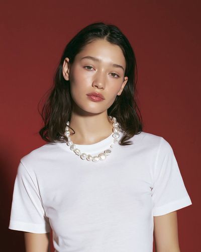 A woman with dark wavy hair wears a pearl necklace in a red studio