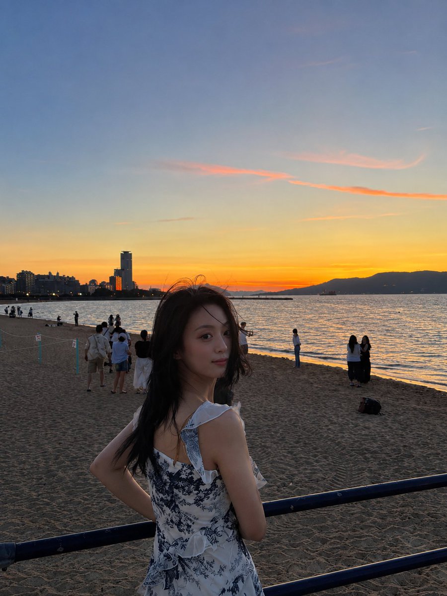 A woman in a blue floral dress looks back at a sunset beach.
